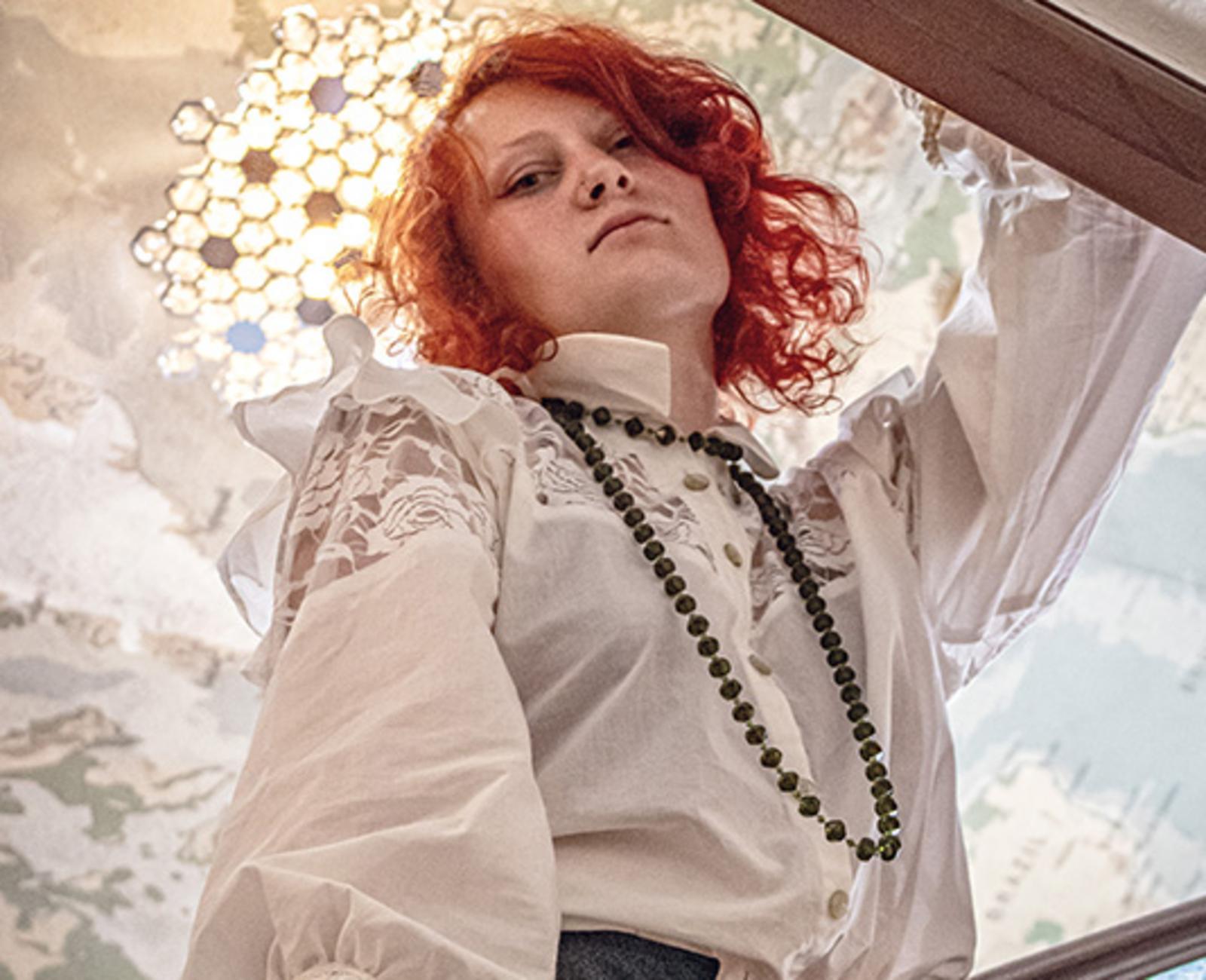 Person mit rotem Haar in weißer Bluse und langer Kette blickt selbstbewusst in die Kamera