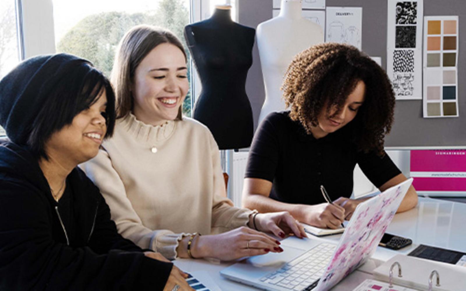 Drei Studierende arbeiten gemeinsam an Laptop und Skizzen im Moderaum