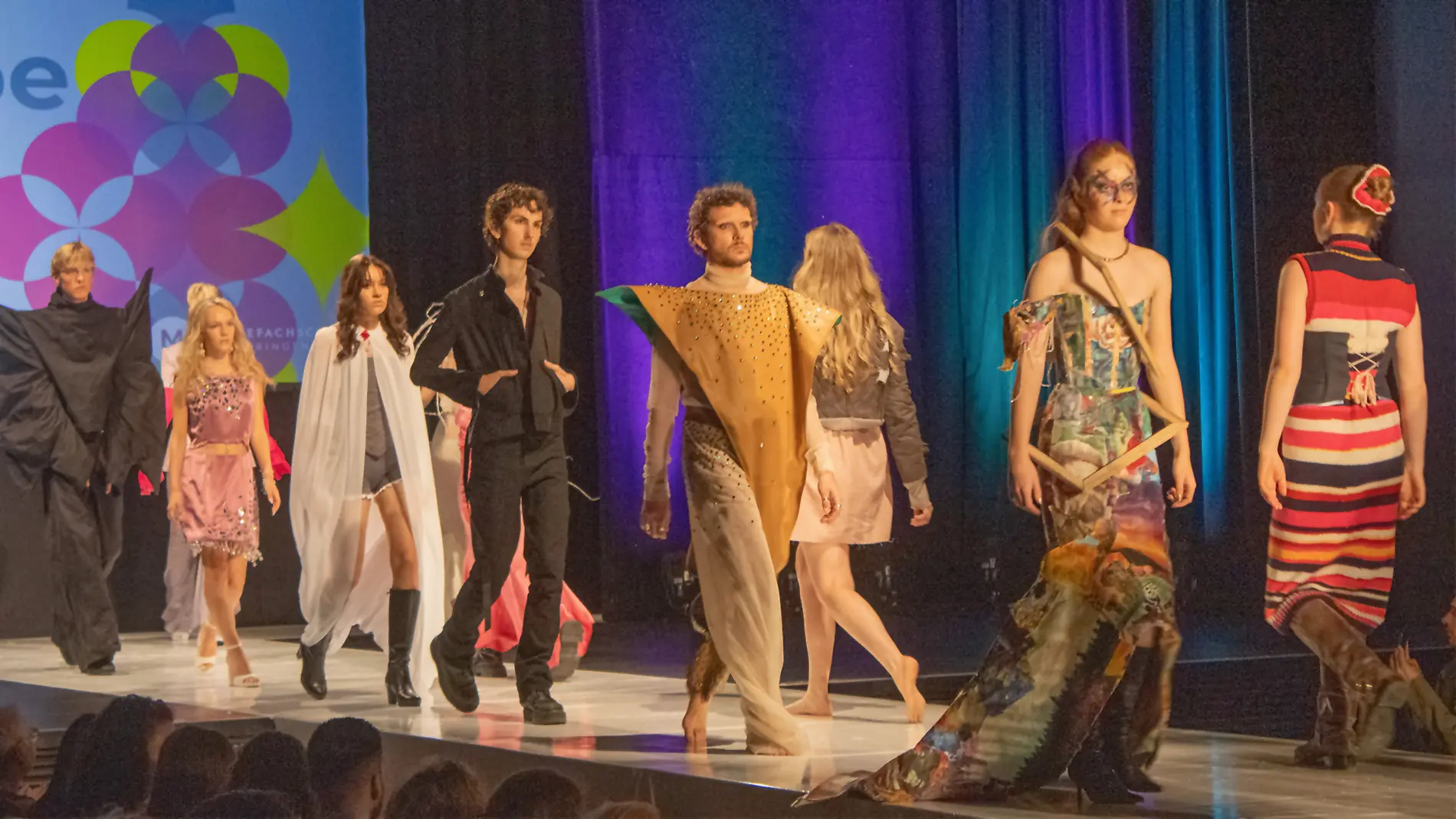Mehrere Models präsentieren kreative und auffällige Outfits auf dem Laufsteg bei einer Modenschau der Modefachschule Sigmaringen. Der Hintergrund ist farbig beleuchtet mit einem Event-Logo.